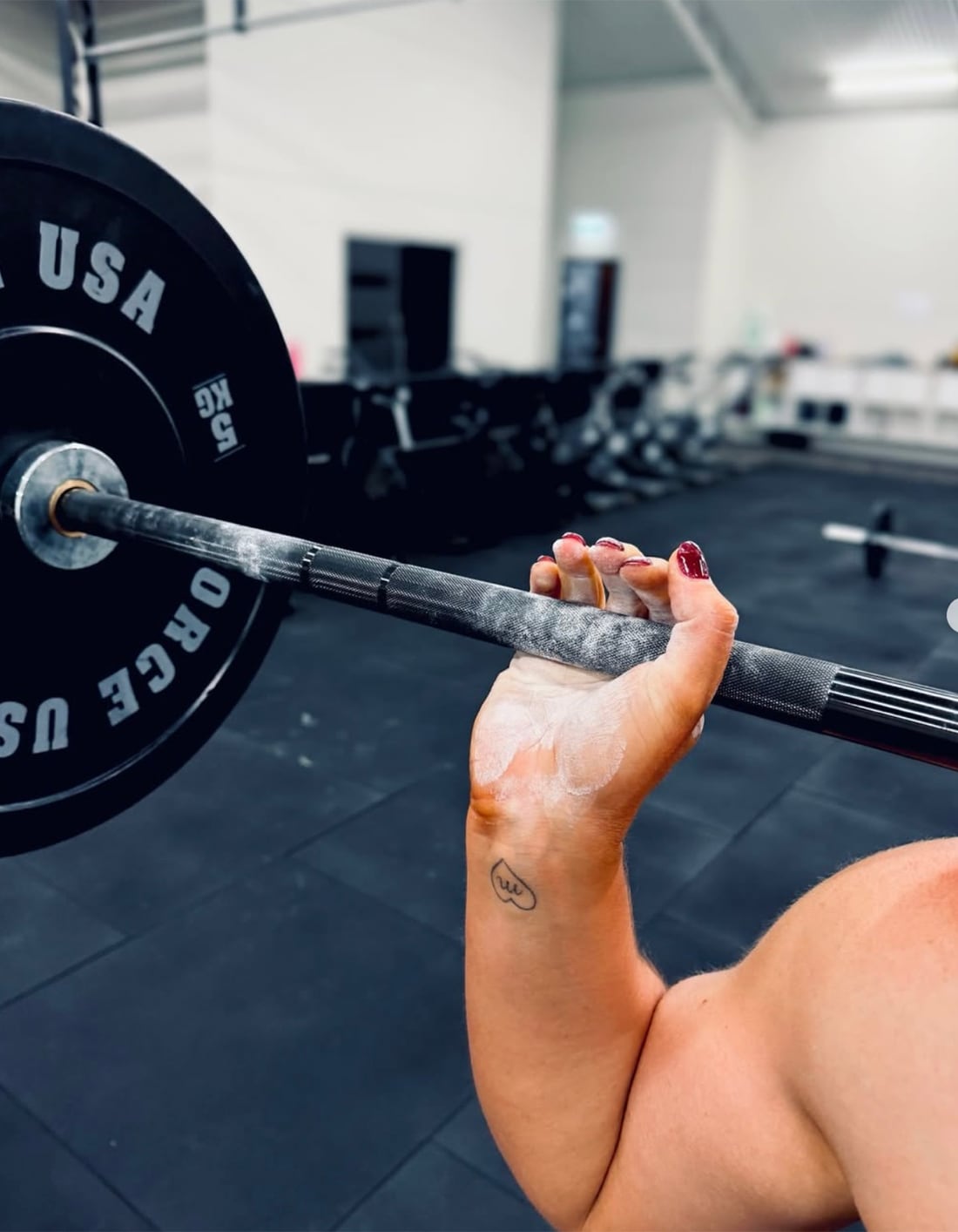 A hand chalked white grips a barbell on the gym floor, dark red nails, a small heart tattoo on the wrist, bumper plate behind.